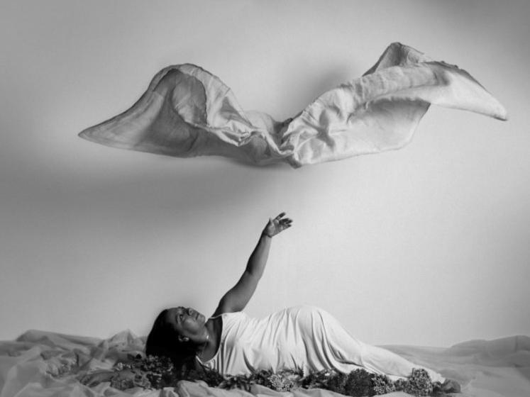 Composición en tonos grises en que aparece una mujer vestida de blanco, acostada en un manto de flores y telas. La mujer con su brazo elevado al cielo, observa una silueta de tela que podría interpretarse como una nube.  