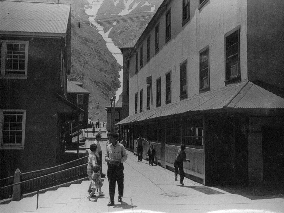 Fotografía en blanco y negro que muestra personas caminando por una ciudad con edificios altos, y de fondo montañas con nieve.